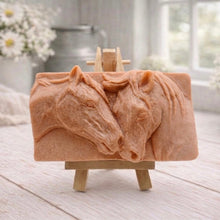 Load image into Gallery viewer, A rectangular handmade vegan soap featuring a detailed relief of two horse heads leaning affectionately towards each other, finished in soft natural tones and displayed on a miniature wooden easel.