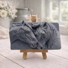 Load image into Gallery viewer, A rectangular handmade vegan soap featuring a detailed relief of two horse heads leaning affectionately towards each other, finished in soft natural tones and displayed on a miniature wooden easel.