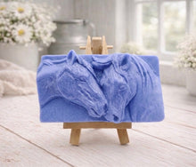 Load image into Gallery viewer, A rectangular handmade vegan soap featuring a detailed relief of two horse heads leaning affectionately towards each other, finished in soft natural tones and displayed on a miniature wooden easel.