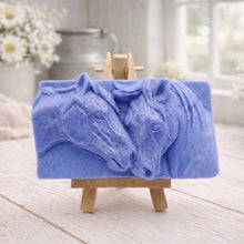 Load image into Gallery viewer, A rectangular handmade vegan soap featuring a detailed relief of two horse heads leaning affectionately towards each other, finished in soft natural tones and displayed on a miniature wooden easel.
