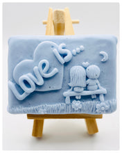 Load image into Gallery viewer, Handmade vegan soaps featuring a boy and girl sat on a bench with the wording Love Is... , in beautiful eco friendly packaging. crafted with natural French clays, activated charcoal and essential oils. a eco-friendly, zero waste gift that’s cruelty-free, skin-nourishing, the perfect gift for Valentines day or anniversary gift. small business UK