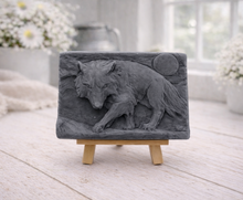 Load image into Gallery viewer, A rectangular handmade vegan soap, featuring a detailed relief carving of a wolf in mid-stride across a rugged landscape with a large full moon in the background, displayed on a miniature wooden easel.