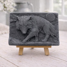 Load image into Gallery viewer, A rectangular handmade vegan soap, featuring a detailed relief carving of a wolf in mid-stride across a rugged landscape with a large full moon in the background, displayed on a miniature wooden easel.