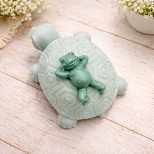 Load image into Gallery viewer, A 3D handmade vegan soap in a soft mint green tone, sculpted into a large friendly turtle with a geometric shell pattern and a small, cute frog sleeping peacefully on its back.