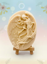Load image into Gallery viewer, Handmade vegan Guardian Angel soap bar in a peach-toned French clay, featuring a detailed angel relief with flowing wings and flowers. Displayed on a small wooden easel. Natural, cruelty-free UK soap ideal for spiritual gifts, zero waste skincare, and sustainable bathroom décor. British-made artisan soap perfect for eco-conscious shoppers