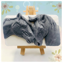 Load image into Gallery viewer, A grey and white handmade vegan soap featuring a highly detailed relief of two horses nuzzling heads, displayed on a wooden easel against a whimsical farmhouse decor background.