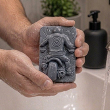 Load image into Gallery viewer, Person holding a bar of soap with decorative detailed handmade vegan soap featuring a motorcycle rider with a skull emblem on their jacket, displayed on a wooden easel against a whimsical farmhouse decor background