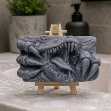 Load image into Gallery viewer, A rectangular handmade vegan soap in a charcoal-grey tone, featuring a high-relief carving of a Tyrannosaurus Rex with its jaws wide open in a roar and sharp claws framing the face, displayed on a miniature wooden easel.