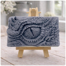 Load image into Gallery viewer, A rectangular handmade vegan soap in a slate-grey tone, featuring a highly detailed, close-up relief of a mythical dragon or dinosaur eye surrounded by thick, pebbled scales and reptilian skin textures, displayed on a miniature wooden easel.