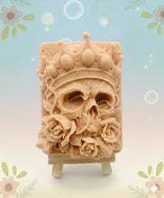 Load image into Gallery viewer, A handmade vegan soap featuring a highly detailed 3D relief of a skull wearing a jewelled crown and surrounded by roses, displayed on a wooden easel against a whimsical background.
