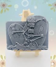 Load image into Gallery viewer, A spooky handmade vegan soap featuring a 3D relief of a skeleton relaxing against "RIP" tombstones, displayed on a wooden easel against a whimsical farmhouse decor background.