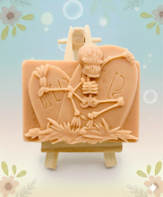 Load image into Gallery viewer, A spooky handmade vegan soap featuring a 3D relief of a skeleton relaxing against "RIP" tombstones, displayed on a wooden easel against a whimsical farmhouse decor background.