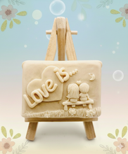 Load image into Gallery viewer, Handmade vegan soaps featuring a boy and girl sat on a bench with the wording Love Is... , in beautiful eco friendly packaging. crafted with natural French clays, activated charcoal and essential oils. a eco-friendly, zero waste gift that’s cruelty-free, skin-nourishing, the perfect gift for Valentines day or anniversary gift. small business UK