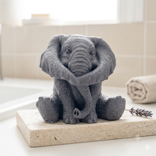Load image into Gallery viewer, A highly detailed handmade vegan soap in a charcoal grey tone, sculpted in the shape of a sitting elephant with realistic wrinkled skin textures and large ears, displayed on a light wooden surface with white flowers in the background.