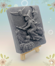 Load image into Gallery viewer, A beautiful handmade vegan soap featuring a detailed image of a witch flying on a broomstick past a crescent moon and owls, displayed on a wooden easel against a whimsical farmhouse decor background.
