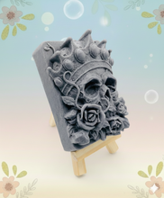 Load image into Gallery viewer, A handmade vegan soap featuring a highly detailed 3D relief of a skull wearing a jewelled crown and surrounded by roses, displayed on a wooden easel against a whimsical background.