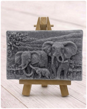 Load image into Gallery viewer, A rectangular handmade vegan soap in a warm grey tone, featuring a detailed relief carving of an elephant family two adults and a small calf walking through a textured savannah landscape, displayed on a miniature wooden easel.