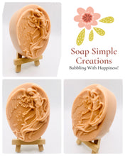 Load image into Gallery viewer, Handmade vegan Guardian Angel soap bar in a peach-toned French clay, featuring a detailed angel relief with flowing wings and flowers. Displayed on a small wooden easel. Natural, cruelty-free UK soap ideal for spiritual gifts, zero waste skincare, and sustainable bathroom décor. British-made artisan soap perfect for eco-conscious shoppers