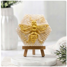 Load image into Gallery viewer, A hexagonal handmade vegan soap featuring a detailed relief of a large queen bee with outstretched wings, highlighted in shimmering gold, set against a textured white honeycomb background and displayed on a miniature wooden easel.