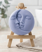 Load image into Gallery viewer, handmade vegan soap gift featuring a circular relief sculpture of a crescent moon and sun with peaceful intertwined faces, displayed on a small wooden easel in a bright farmhouse decor setting.