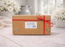 Load image into Gallery viewer, Brown gift box with a red ribbon on a wooden surface with blurred flowers in the background