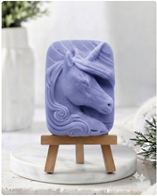 Load image into Gallery viewer, handmade vegan soap featuring a detailed unicorn head relief with a spiralled horn and flowing curly mane, displayed on a small wooden easel in a bright farmhouse decor setting.