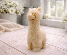 Load image into Gallery viewer, handmade vegan soap in the shape of a realistic standing alpaca sculpture, featuring detailed curly wool texture and a smiling face, displayed on a rustic white wooden surface in a bright farmhouse decor setting.