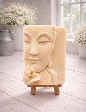 Load image into Gallery viewer, handmade vegan soap featuring a serene Buddha face relief with a lotus flower, displayed on a small wooden easel in a bright farmhouse decor setting with flowers and a rustic milk churn.