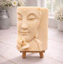 Load image into Gallery viewer, handmade vegan soap featuring a serene Buddha face relief with a lotus flower, displayed on a small wooden easel in a bright farmhouse decor setting with flowers and a rustic milk churn.
