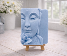 Load image into Gallery viewer, handmade vegan soap featuring a serene Buddha face relief with a lotus flower, displayed on a small wooden easel in a bright farmhouse decor setting with flowers and a rustic milk churn.