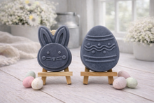 Load image into Gallery viewer, handmade vegan soap gift set featuring a cute bunny face and a decorated Easter egg with wave patterns, displayed on wooden easels against a rustic white farmhouse decor background with spring flowers.