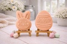 Load image into Gallery viewer, handmade vegan soap gift set featuring a cute bunny face and a decorated Easter egg with wave patterns, displayed on wooden easels against a rustic white farmhouse decor background with spring flowers.