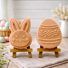 Load image into Gallery viewer, handmade vegan soap gift set featuring a cute bunny face and a decorated Easter egg with wave patterns, displayed on wooden easels against a rustic white farmhouse decor background with spring flowers.