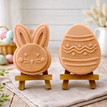 Load image into Gallery viewer, handmade vegan soap gift set featuring a cute bunny face and a decorated Easter egg with wave patterns, displayed on wooden easels against a rustic white farmhouse decor background with spring flowers.