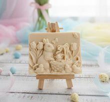 Load image into Gallery viewer, handmade vegan soap featuring a highly detailed 3D relief of a rabbit in a garden with tulips and an Easter basket, displayed on a wooden easel in a rustic farmhouse decor setting.