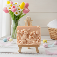 Load image into Gallery viewer, handmade vegan soap featuring a highly detailed 3D relief of a rabbit in a garden with tulips and an Easter basket, displayed on a wooden easel in a rustic farmhouse decor setting.