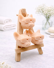 Load image into Gallery viewer, A set of three handmade vegan soaps in the shape of French Bulldog heads wearing sunglasses, displayed on a wooden easel in a bright, modern bathroom setting.
