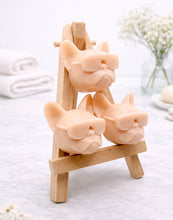 Load image into Gallery viewer, A set of three handmade vegan soaps in the shape of French Bulldog heads wearing sunglasses, displayed on a wooden easel in a bright, modern bathroom setting.