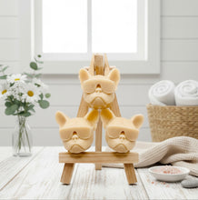 Load image into Gallery viewer, A set of three handmade vegan soaps in the shape of French Bulldog heads wearing sunglasses, displayed on a wooden easel in a bright, modern bathroom setting.