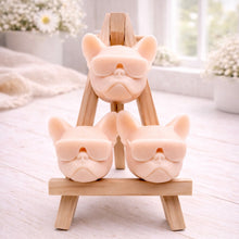 Load image into Gallery viewer, A set of three handmade vegan soaps in the shape of French Bulldog heads wearing sunglasses, displayed on a wooden easel in a bright, modern bathroom setting.