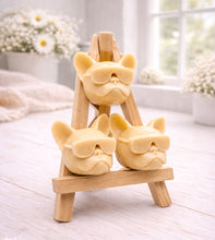 Load image into Gallery viewer, A set of three handmade vegan soaps in the shape of French Bulldog heads wearing sunglasses, displayed on a wooden easel in a bright, modern bathroom setting.