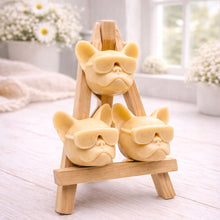 Load image into Gallery viewer, A set of three handmade vegan soaps in the shape of French Bulldog heads wearing sunglasses, displayed on a wooden easel in a bright, modern bathroom setting.