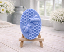 Load image into Gallery viewer, handmade vegan soap featuring an oval relief sculpture of two honey bees crawling on a honeycomb pattern, displayed on a small wooden easel in a bright farmhouse decor setting.