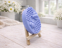 Load image into Gallery viewer, handmade vegan soap featuring an oval relief sculpture of two honey bees crawling on a honeycomb pattern, displayed on a small wooden easel in a bright farmhouse decor setting.