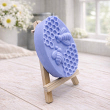 Load image into Gallery viewer, handmade vegan soap featuring an oval relief sculpture of two honey bees crawling on a honeycomb pattern, displayed on a small wooden easel in a bright farmhouse decor setting.
