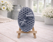 Load image into Gallery viewer, handmade vegan soap featuring an oval relief sculpture of two honey bees crawling on a honeycomb pattern, displayed on a small wooden easel in a bright farmhouse decor setting.