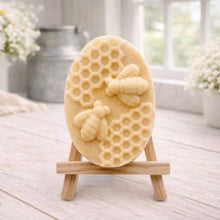 Load image into Gallery viewer, handmade vegan soap featuring an oval relief sculpture of two honey bees crawling on a honeycomb pattern, displayed on a small wooden easel in a bright farmhouse decor setting.