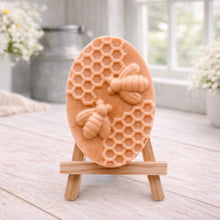 Load image into Gallery viewer, handmade vegan soap featuring an oval relief sculpture of two honey bees crawling on a honeycomb pattern, displayed on a small wooden easel in a bright farmhouse decor setting.