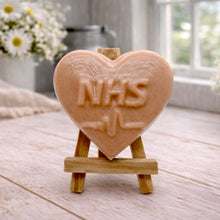 Load image into Gallery viewer, handmade vegan soap in a heart shape featuring an "NHS" relief and a heartbeat pulse line, displayed on a small wooden easel in a bright farmhouse decor setting.