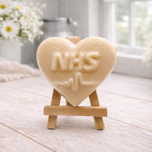 Load image into Gallery viewer, handmade vegan soap in a heart shape featuring an "NHS" relief and a heartbeat pulse line, displayed on a small wooden easel in a bright farmhouse decor setting.